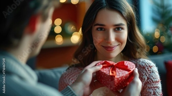 Fototapeta Young man is giving a present to his lovely girlfriend while they are sitting on couch at home