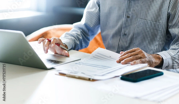 Fototapeta Asian businessman using computer laptop working and reviewing document reports at office workplace. legal expert, professional lawyer reading and checking financial documents or insurance contract