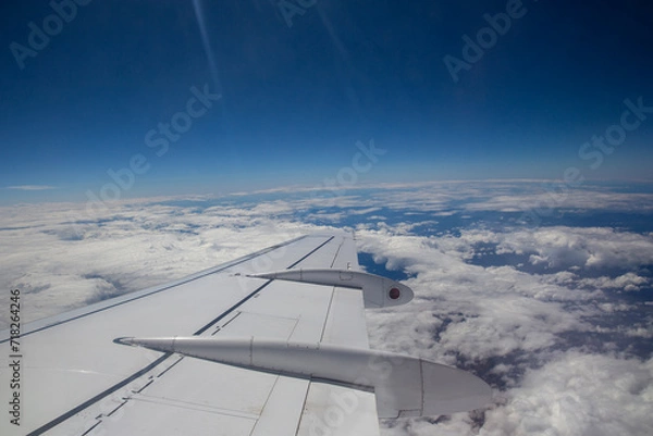 Fototapeta Aerial view by the airplane wing while flying above the clouds with a blue sky in the background, wide angle shot