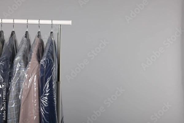 Fototapeta Dry-cleaning service. Many different clothes in plastic bags hanging on rack against grey background, space for text