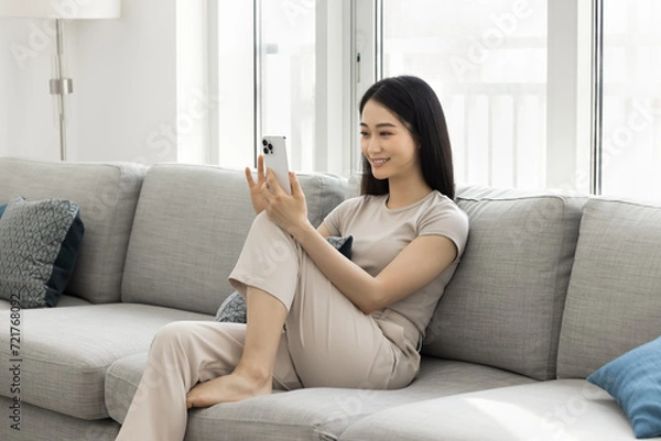 Fototapeta Happy beautiful young Asian blogger woman recording self video, talking selfie, talking on conference call on smartphone, using mobile phone on home sofa, holding gadget with knee support