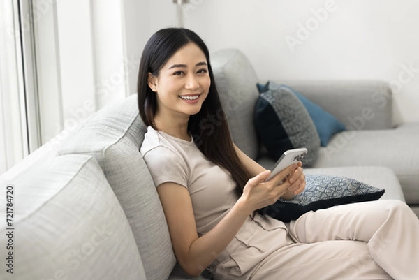 Fototapeta Happy beautiful young Asian customer girl using mobile phone at home, promoting new online application, Internet service, e-commerce platform, smart home app, looking at camera, smiling