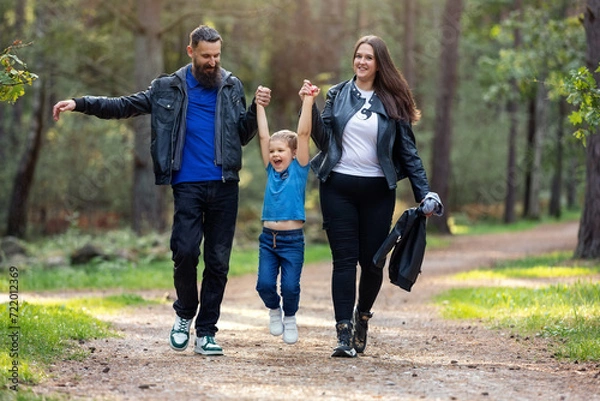 Fototapeta Mother and father lift their son up by the arms during a walk in the forest path. An active child likes such a game, he has fun. Nice day, happy family