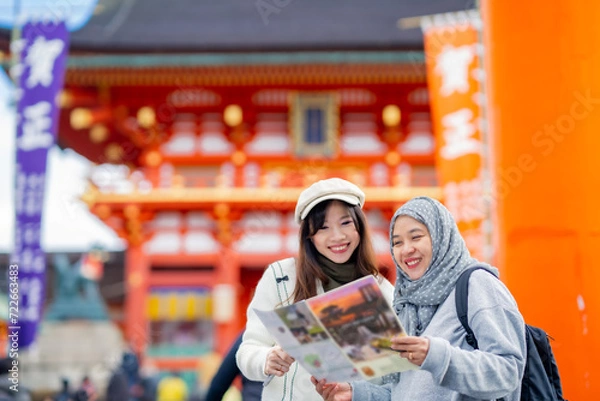 Obraz Travel, muslim, Two Asian female tourists of different religions friends visitor learning about history of fushimi inari shrine in travel book while walking through senbon torii path in Kyoto Japan.