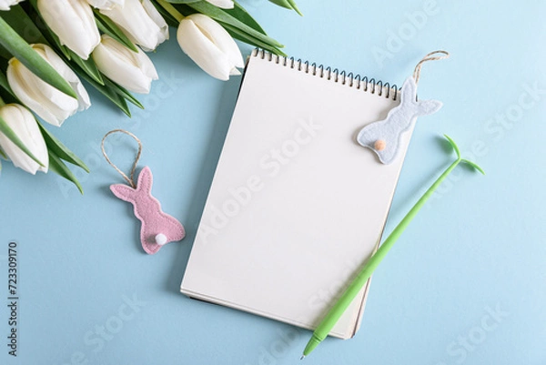 Fototapeta Floral border from bouquet of white tulips, Easter eggs and blank notebook on blue background. Easter celebration concept. Copy space. Top view