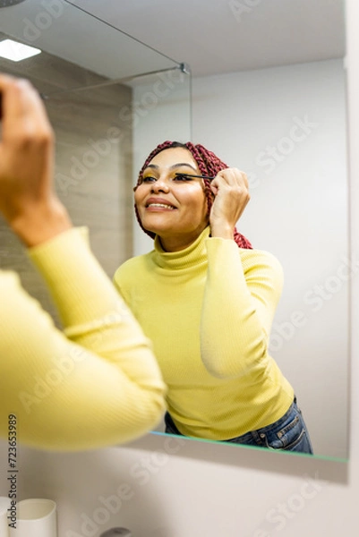 Fototapeta Vertical photo of a dark-skinned woman using mascara to enhance her eyelashes in front of the bathroom mirror. Concept of African women putting on makeup in front of the mirror.