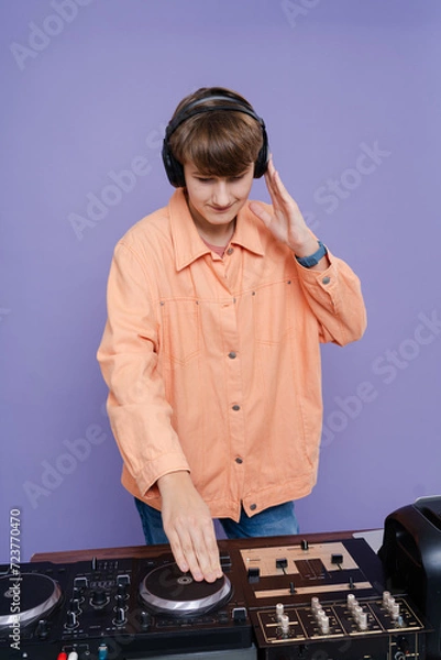 Fototapeta Cool young boy DJ mixes music on a mixing console  in stylish clothes on a color background.