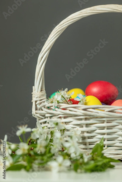 Fototapeta white wicker basket with colored eggs and spring flowers on the table, happy Easter concept