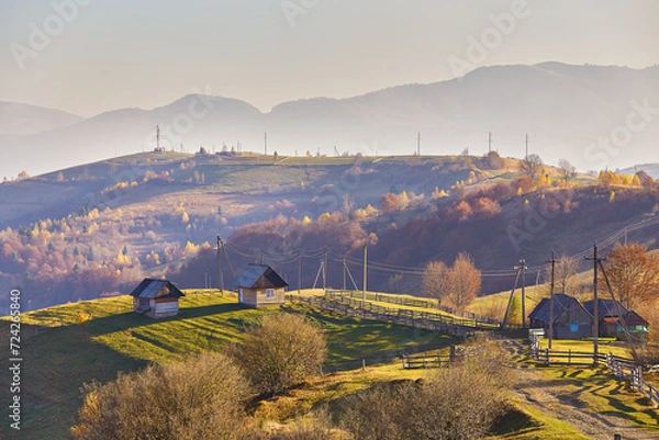 Fototapeta Panoramic view of misty sunny autumn evening in mountains. Beautiful sunset hills landscape. Slopes, meadows, fields, village, house, dirt road. Amazing fall rural scene. Carpathian range.