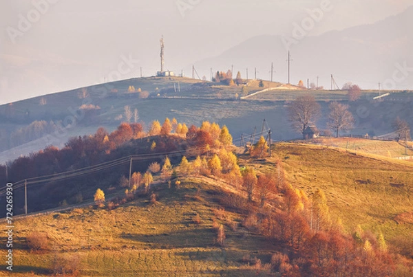 Fototapeta Panoramic view of misty sunny autumn evening in mountains. Beautiful sunset hills landscape. Slopes, meadows, fields, village, house, dirt road. Amazing fall rural scene. Carpathian range.
