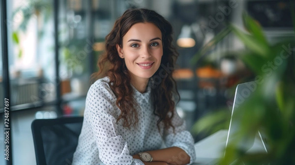 Fototapeta Smiling young woman with curly hair at her computer in a modern office with greenery