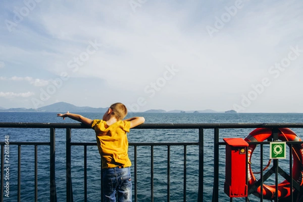 Fototapeta A boy in a yellow T-shirt stands on the deck of the ferry and looks at the sea. The child looks into the distance