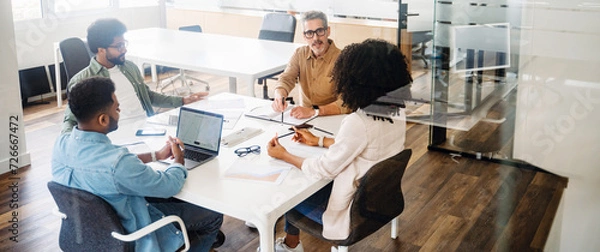 Fototapeta In a wide office shot, four professionals are deep in discussion around a table, showcasing a dynamic and collaborative team in action. A diverse team in a friendly and inclusive workplace atmosphere