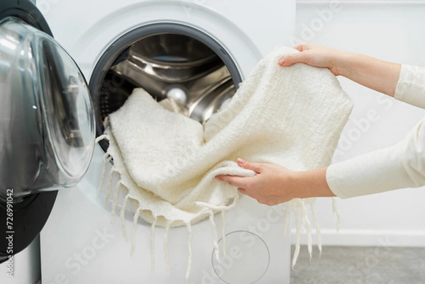 Fototapeta Woman unloading laundry, taking white wool blanket out of the drum of washing machine in laundry room