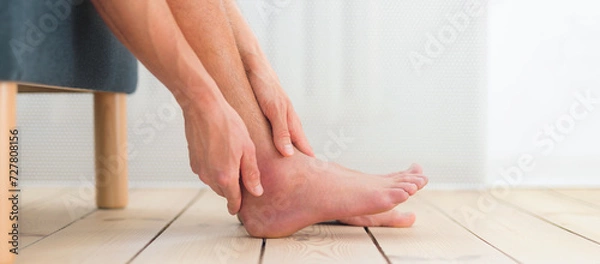 Obraz Ankle sprain. Man hold his sprained, swollen ankle sitting on the sofa at home.