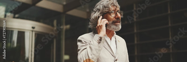 Fototapeta Mature businessman with beard and glasses wearing gray jacket walks up the stairs leaving the business center, Panorama. Middle aged manager talking on cellphone
