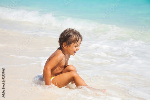 Fototapeta Happy boy sits on the sand beach with white waves