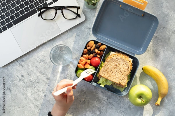 Fototapeta Healthy business lunch at workplace. Sandwich, vegetables, fruid and water lunch box on working desk with laptop, glasses.