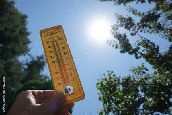Fototapeta Hand holding thermometer towards the sun and clear sky showing representing raising temperatures and hot weather