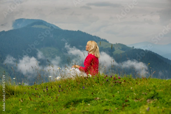 Fototapeta A middle-aged blonde woman sits in a clearing high in the mountains among wild flowers and clouds. A moment of pleasure.