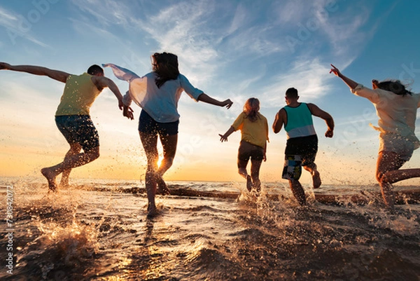 Obraz Group of happy young friends are having fun, runs and jumps at sunset beach with lots of splashes