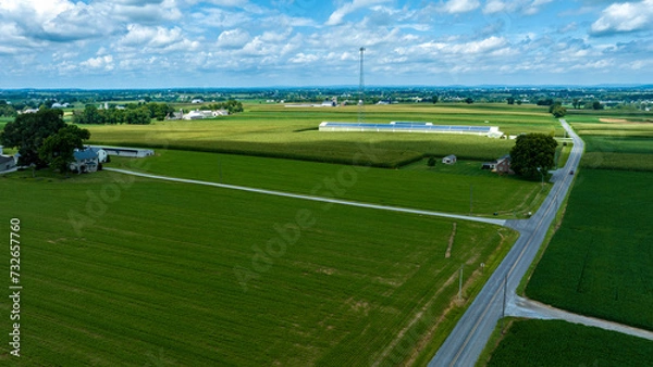 Fototapeta Aerial View Of A Lush Green Farmland With A Crossroad, Adjacent Fields Of Different Crops, And A Farmhouse Next To A Large Building With Solar Panels On Its Roof Under A Partly Cloudy Sky.