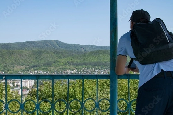 Fototapeta A man in a cap with a backpack stands on the observation deck and looks at the city and the mountains