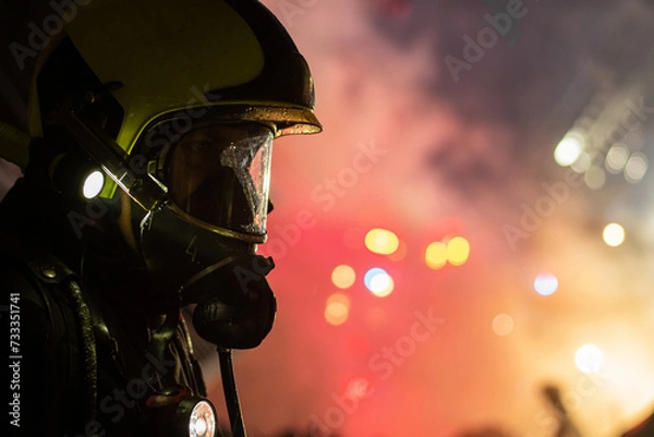 Fototapeta Close-up of a firefighter wearing a helmet and breathing apparatus mask at a night fire with blue and red lights in the background