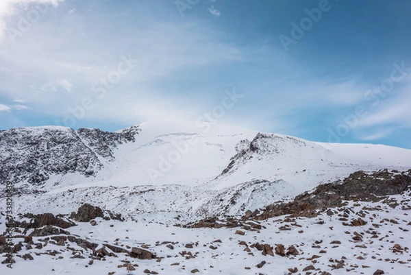 Fototapeta Cold snow-white landscape with snow-capped peak of dome shape under clouds in blue sky. Snow-covered rocky mountain range with snowy dome shaped mountain top. Pure white snow and stones in mountains.