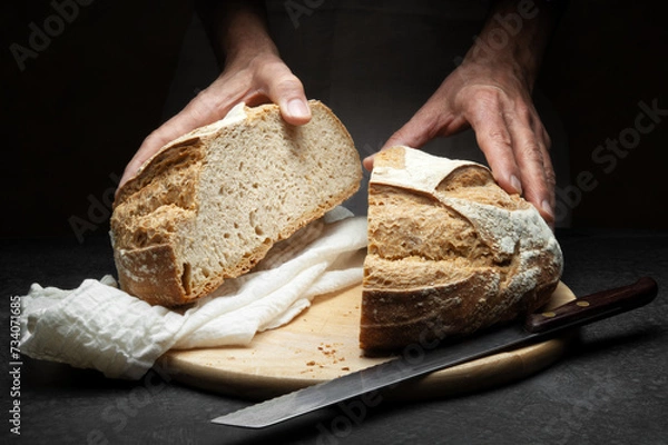 Fototapeta Hands with loaf of bread cut in half on worktop with cutting board, knife and tea towel, close-up.