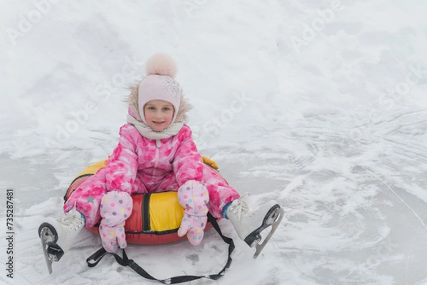 Fototapeta A little girl of 5 years old sits on a tubing and relaxes after skating on white figure skates at the skating rink in winter.