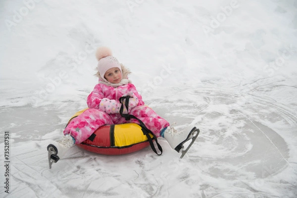 Fototapeta A little girl of 5 years old sits on a tubing and relaxes after skating on white figure skates at the skating rink in winter.
