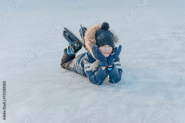 Fototapeta 7 year old boy lying on the ice, skates on his feet, resting from skating. Winter fun.