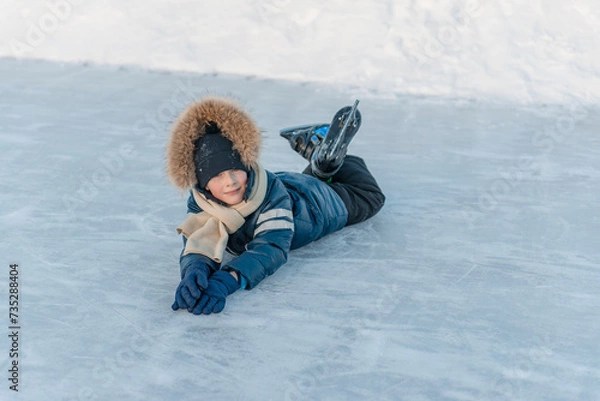Fototapeta 7 year old boy lying on the ice, skates on his feet, resting from skating. Winter fun.