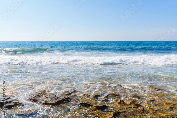 Fototapeta Enchanting Calm Blue Ocean Waves Hitting and Splashing on the Seashore and Beach Under the Clear Sky, During a Hot Sunny Summer Day Morning 