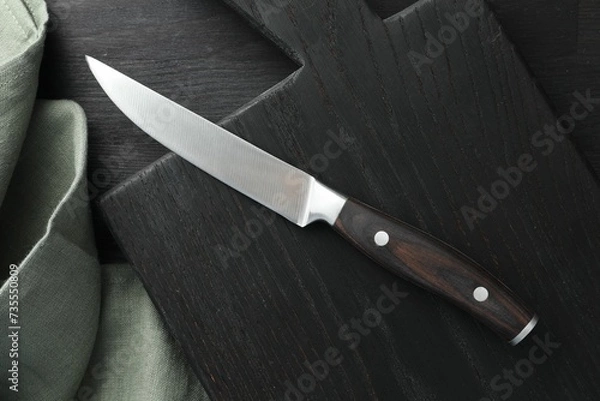 Fototapeta One sharp knife, cloth and board on black wooden table, top view