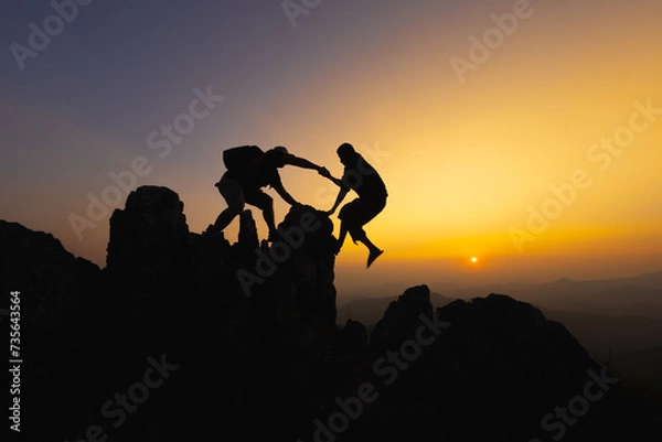 Fototapeta  Silhouettes of two people climbing on mountain and helping. Teamwork of two men hiker helping each other on top of mountain climbing team beautiful sunrise. Help and assistance concept.