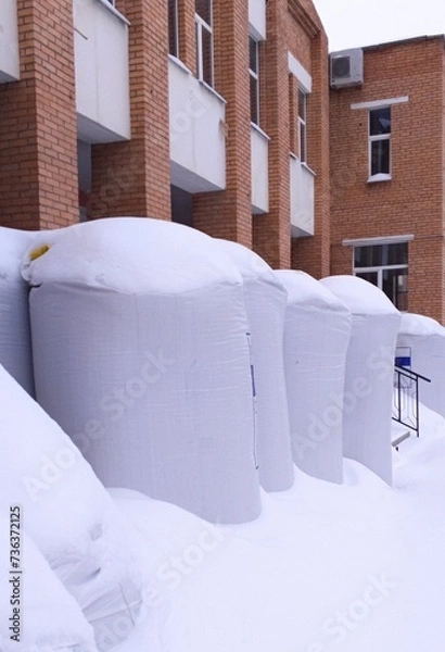 Fototapeta Large bags of expanded clay stand near a brick building and are covered with snow