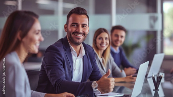 Fototapeta A confident professional man in a suit is giving a thumbs up in a bright office environment with cheerful colleagues around him, indicating success and approval.
