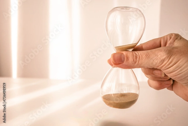Obraz woman's hand holding time, depicted on an elegant glass hourglass with morning light background