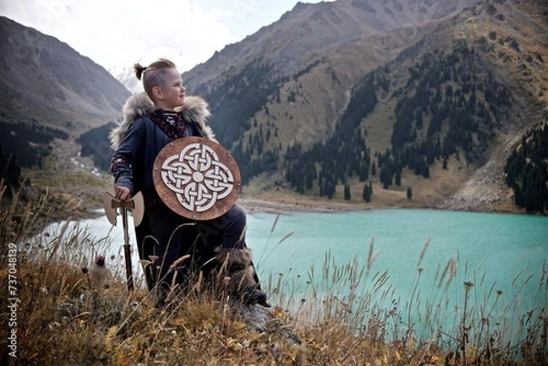 Fototapeta A young boy in the viking cosplay costume holding a wooden shield and a axe and standing next to the beautiful alpine lake with Mountains view