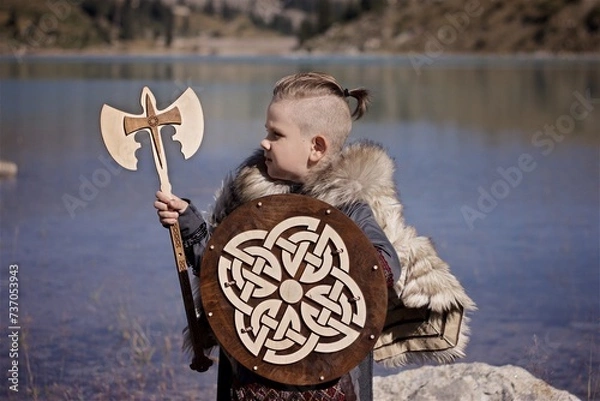 Fototapeta A young boy in the viking cosplay costume holding a wooden shield and a axe and standing next to the beautiful alpine lake with Mountains view