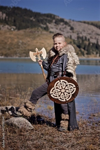 Fototapeta A young boy in the viking cosplay costume holding a wooden shield and a axe and standing next to the beautiful alpine lake with Mountains view