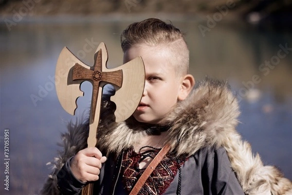 Fototapeta Portrait of a young boy in the viking cosplay costume holding a wooden shield and a axe and standing next to the beautiful alpine lake with Mountains view