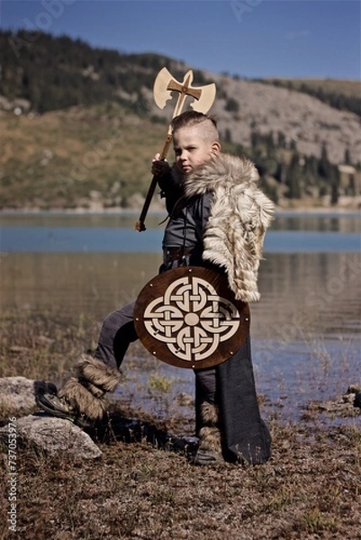 Fototapeta A young boy in the viking cosplay costume holding a wooden shield and a axe and standing next to the beautiful alpine lake with Mountains view