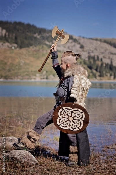 Fototapeta A young boy in the viking cosplay costume holding a wooden shield and a axe and standing next to the beautiful alpine lake with Mountains view
