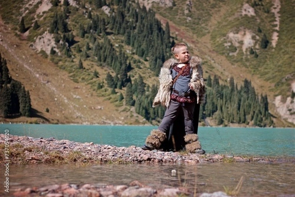Fototapeta A young boy in the viking cosplay costume standing next to the beautiful alpine lake with Mountains view