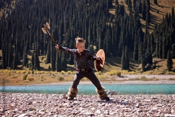 Fototapeta A young boy in the viking cosplay costume holding a wooden shield and a axe and training next to the beautiful alpine lake with Mountains view