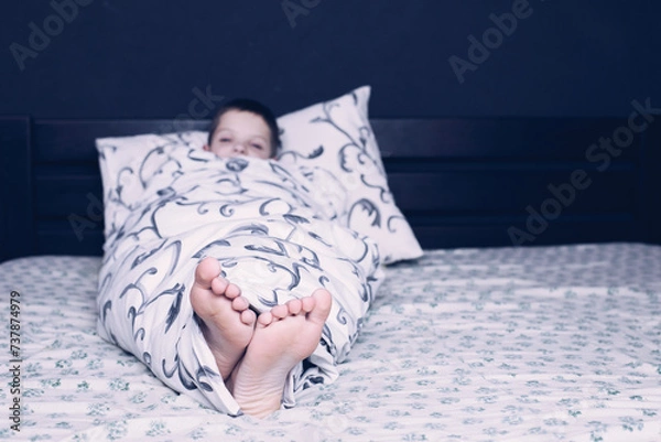 Fototapeta Bare feet of a child. A child in pajamas is smiling. The child is wrapped in a blanket. The boy is sleeping in bed. Foot and leg
