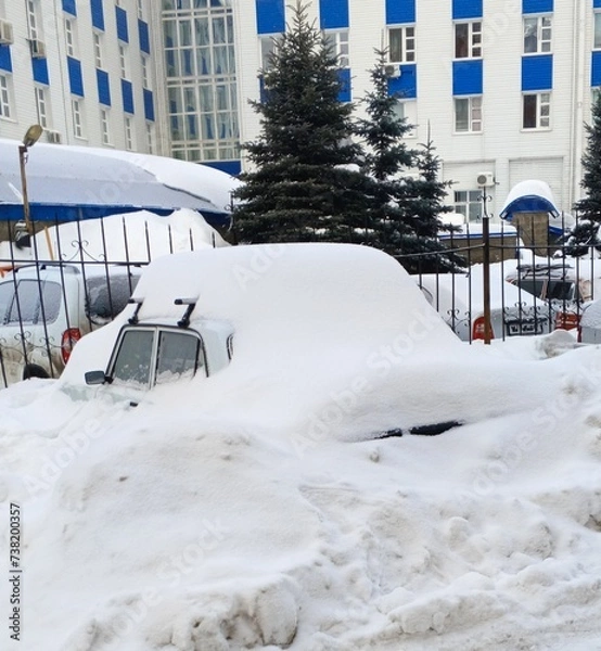 Fototapeta There is a car near the fence, covered with a thick layer of snow against the background of a building and fir trees